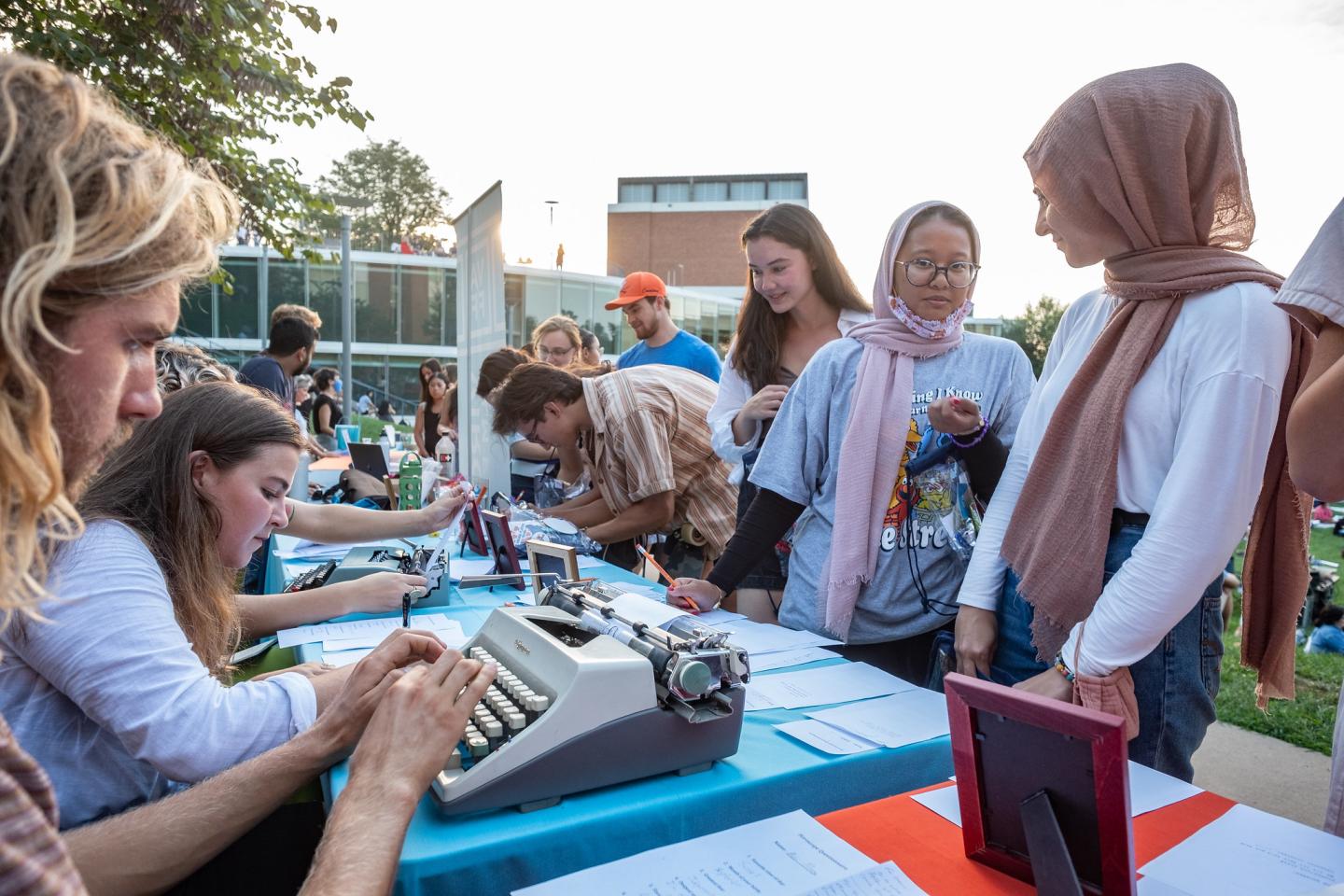 A blonde person types a poem on a typewriter while someone sits next to them. Students stand in front of the table, chatting and watching. 