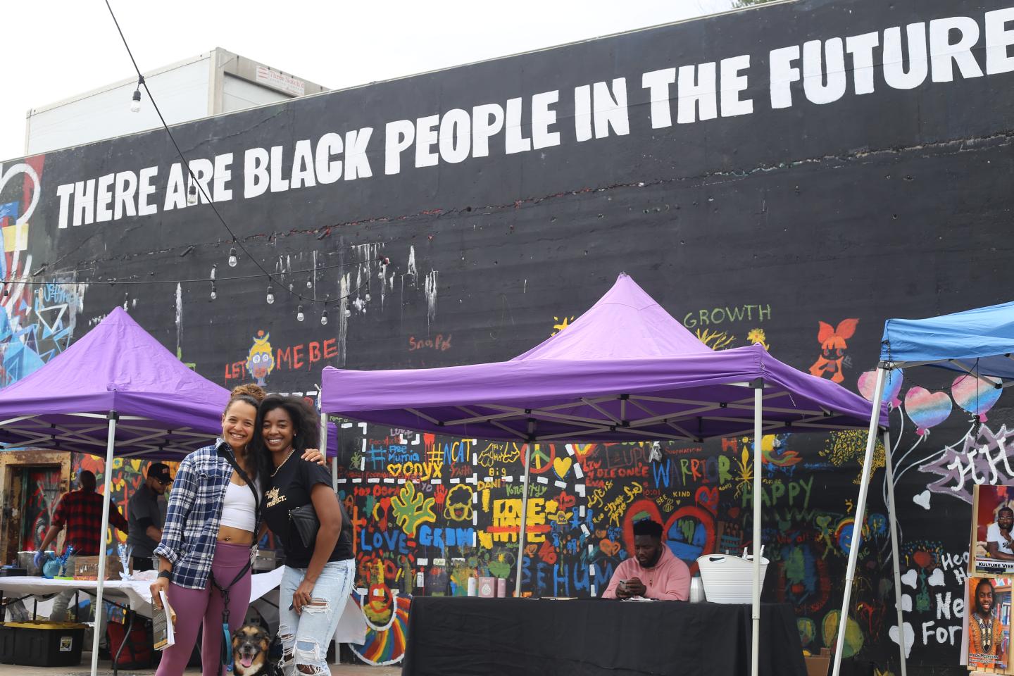 Two people near purple tents with a mural in the background that says "There are Black People in the Future"