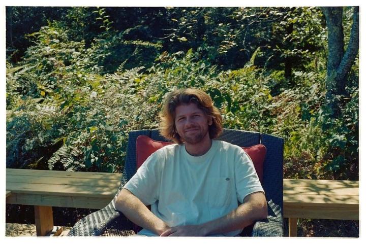 Graham Barbour smiles for the camera wearing a white t-shirt and sitting outdoors.