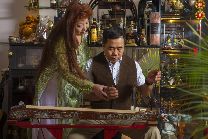 Master Dàn bau player Nam Phuong Nguyen and her apprentice Anh Dien Ky Nguyen. In the background, there is a shelf full of various nicknacks. 